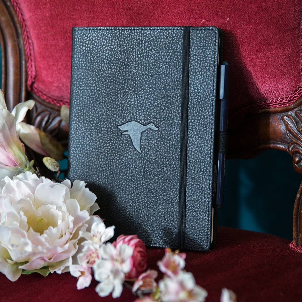 Svart anteckningsbok Wildlife Dotted A5 Black Duck från Dingbats* . Boken står på en stol med röd sammet. Framför boken finns det skira blommor.- Tidformera