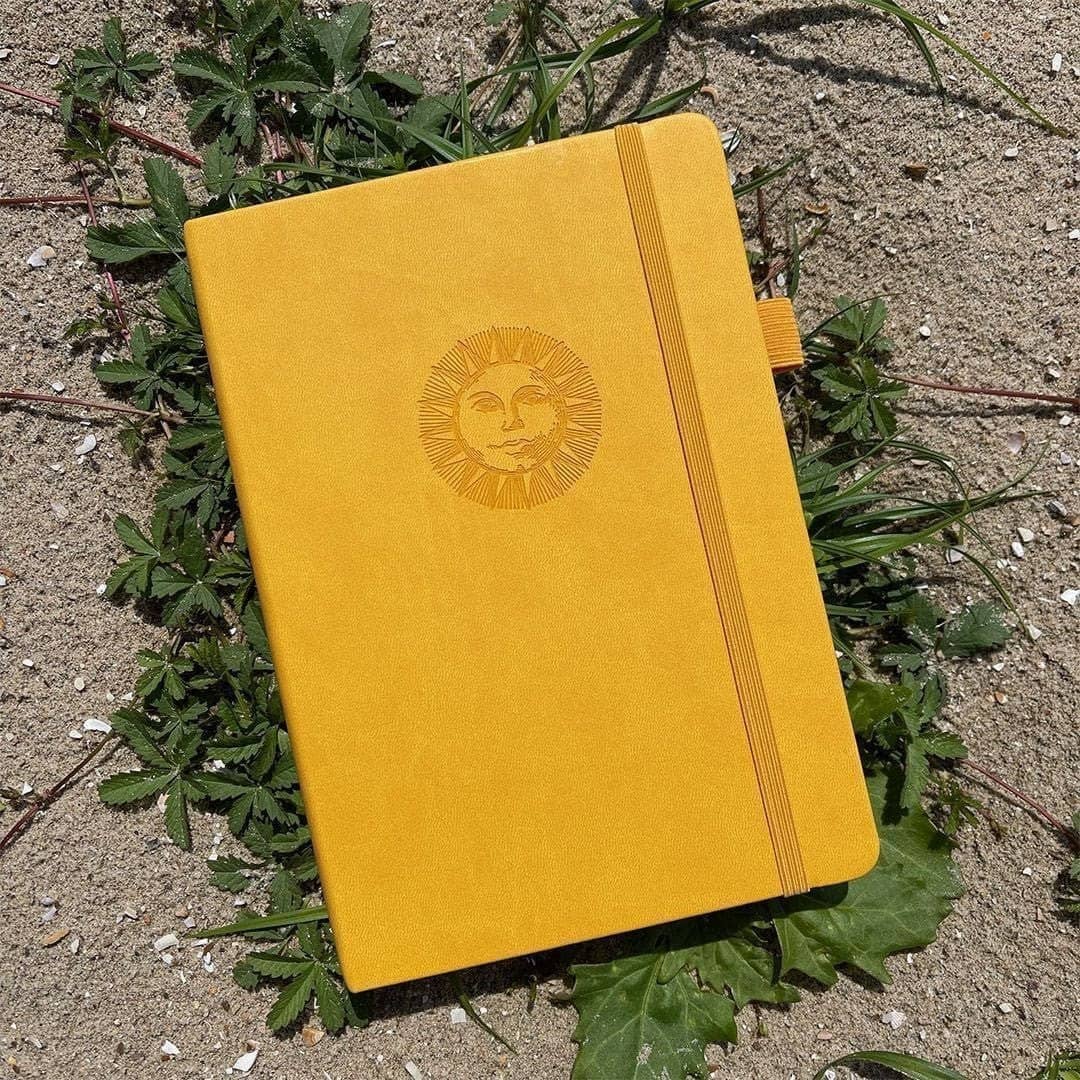 Dotted Notebook A5 160 GSM Smiling sun frÄn Paper24. Gul anteckningsbok med motiv med en leende sol. Skrivboken ligger ute i solen pÄ vÀxter. - Tidformera