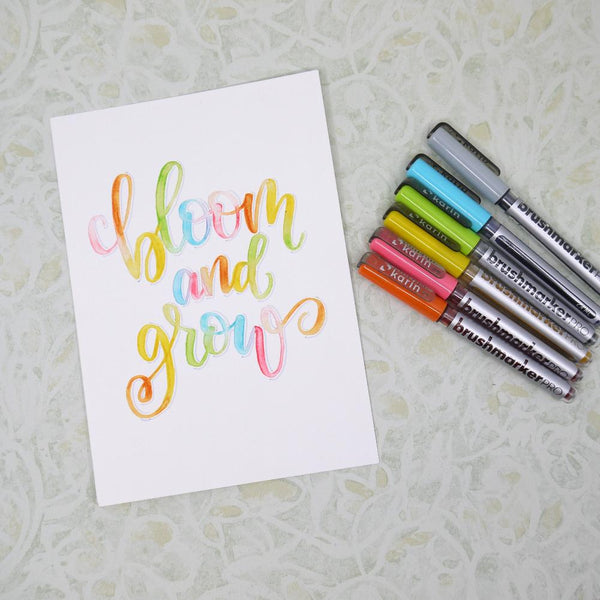 Brushmarker Pro pennor ligger bredvid ett papper med texten Bloom and grow. Texten är skrivet med brush lettering och de pennorna.