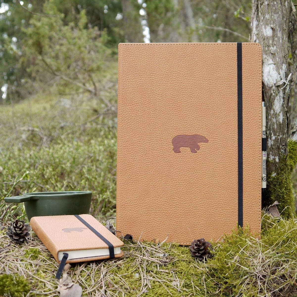 Anteckningsbok Wildlife Dotted A4 Brown Bear från Dingbats*. Två anteckningsböcker, varav en är A4 och den andra är A6 storlek, med det bruna björnmotivet. De finns i skogen bland mossa och kottar. Bakom den lilla boken finns en kåsa. - Tidformera