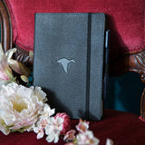 Svart anteckningsbok Wildlife Dotted A5 Black Duck från Dingbats* . Boken står på en stol med röd sammet. Framför  boken finns det skira blommor.- Tidformera