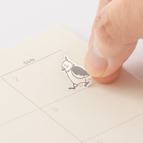 Kalenderstickers Djurtema - Birds från  Midori. Ett finger fäster ett  klistermärke med en fågel i en kalender. Tidformera