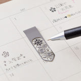 Ett bokmärke ligger på en kalender. En penna ritar i stencilen på bokmärket. - Clip Bookmarker Flower från Midori.