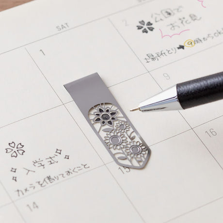 Ett bokmärke ligger på en kalender. En penna ritar i stencilen på bokmärket. - Clip Bookmarker Flower från Midori.